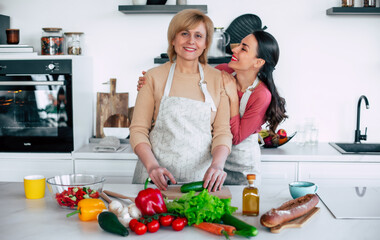 Smiling mature mother and her expressive lovely adult daughter cutting vegetables for a vegan salad together. Mid-adult woman and cute girl are preparing proper healthy meal on domestic kitchen - 777366239
