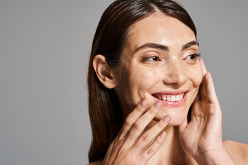 A young Caucasian woman with brunette hair smiles warmly, gently touching her face with her hands...