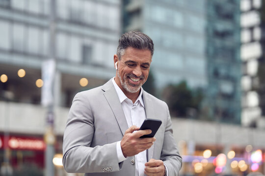 Happy middle aged business man wearing suit earbud holding phone having call walking on city street. Mature businessman with earphone using smartphone listening podcast in app on cellphone outdoors. - Powered by Adobe