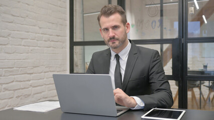 Middle Aged Businessman Looking at Camera while Working on Laptop