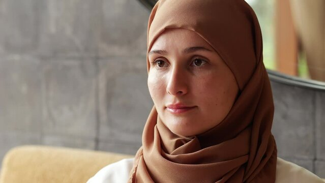 Young adult woman wearing hijab half-side portrait, she sitting and confidently look at something at left. Close headshot with blurred background, natural light from window