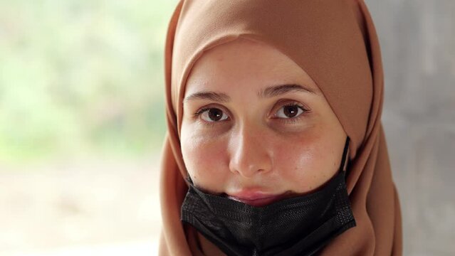 Portrait shot, woman pulls down her medical mask, revealing her face, turns her head and looks directly into the camera. Young lady wearing hijab wrapped around her head and covering hair.