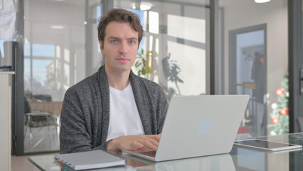 Young Man Looking at Camera while Working on Laptop in Office