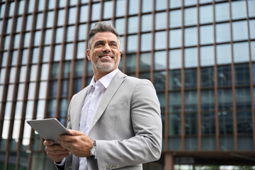 Happy business man investor of middle age in suit holding digital tablet, mature professional businessman leader using tab looking away standing in busy office city street thinking of future success. - 777393640