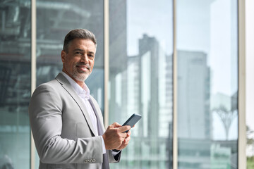 Happy rich middle aged businessman using cellphone, busy older business man leader investor, mature male executive in suit holding mobile phone standing at office window looking at camera, portrait.
