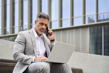 Busy middle aged business man merchant making phone call using laptop sitting outside office. Mature businessman manager wearing suit talking on cellphone working on computer consulting client. - 777399014
