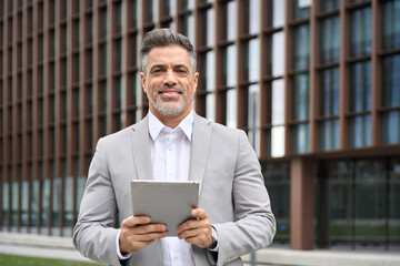 Happy middle aged male entrepreneur, rich older investor, confident business man ceo executive leader, mature businessman manager financial advisor in suit using tablet standing outdoors, portrait. - 777400831