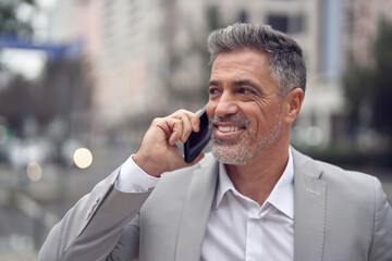 Busy happy middle aged business man making call on mobile phone walking on city street. Smiling mature businessman talking on cellphone standing outside office looking away. Close up. - 777401280