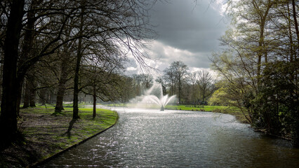 The beautiful city park of the city of Apeldoorn with the large ponds and beautiful fountain on an early spring day with a heavily cloudy sky, province of Gelderland, the Netherlands
