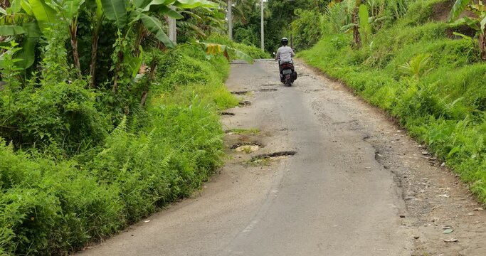 Man rides motorbike on narrow, damaged asphalt road, driving around deep potholes. The sloping carriageway has been eroded by water flows and vehicles