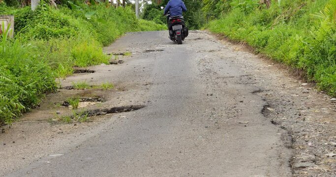 Man ride motorbike on road with many potholes, keep away from them. Very bad asphalt surface condition caused by weathering and water flows due to broken drainage. Village street at central Bali