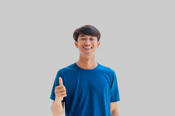 A young Asian man in his 20s wearing a blue t-shirt with a smile pointing thumbs up isolated on a gray background. Well done. Agreement concept. Happy man