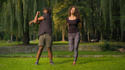 Active healthy couple Indian Arabian man and Caucasian woman athletes doing sport morning workout stretching body muscles warm-up exercises in park outdoors sporty people diverse male female training