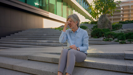 Caucasian tired business woman mature old senior sick unwell businesswoman on city stairs rest...