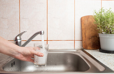 Woman hand holding a clear glass under a stream of fresh tap water from a kitchen sink faucet ,...