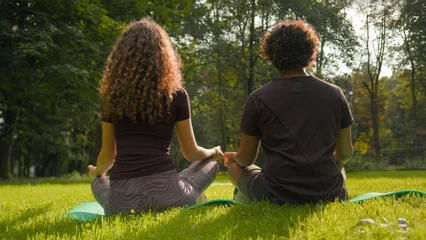 Kissenbezug Back view woman and man doing morning exercise in city park nature green grass fitness couple sit in lotus pose meditate spiritual practice lesson meditation yoga calm mindfulness peace zen relaxation © Yuliia