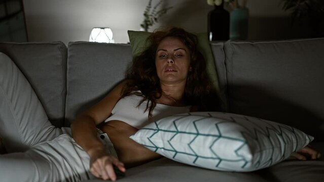A young woman lounges on a couch at home, looking tired or unwell, holding a remote in a dimly lit room.