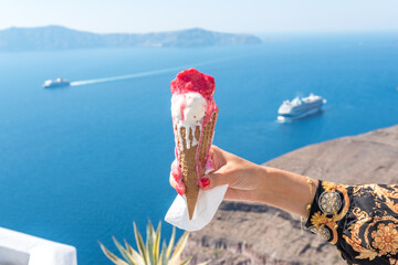 Ice cream in a waffle cone in woman's hand on background of Santorini island and Caldera in Aegean...