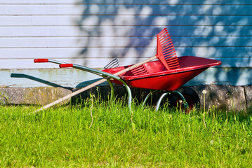 Red common steel wheelbarrow loaded with a red plastic garden rake in front of a white wooden house...