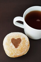 Fluffy donut with chocolate heart next to white cup of coffee on brown background