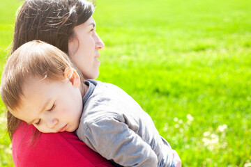 Little boy sleeping on his mother's shoulders next to a green spring meadow.