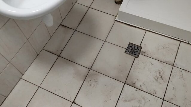 Old dirty tiled bathroom with shower and dirty floor with traces of shoes in a cheap hostel or student dormitory