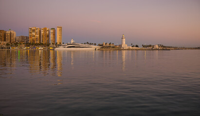 Fototapeta premium Panoramic view with the first lights and its reflections in the water, of lighthouse called La Farola, pier 1 and a yacht. Malaga, Spain
