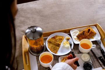 Woman is eating desserts in bed from wooden tray in hotel room on vacations, top view. Pancakes, cheesecakes, pastry with tea and ice-cream, condensed milk, sour cream and jam. Resting and relax.