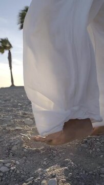 Copy space vertical footage of unrecognisable black woman walking barefoot on the sand beach wearing white trousers that move with the breeze, sunset and palm trees at the background. Summer vibes.