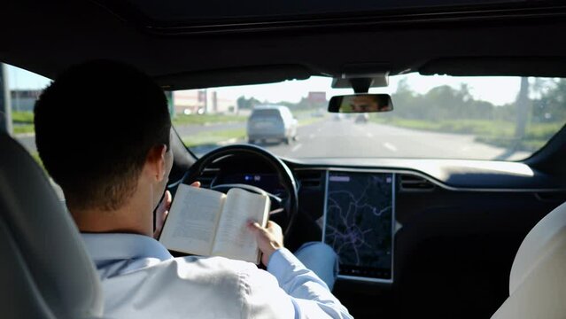 Male businessperson reading book during riding on electrical vehicle with autopilot at urban road. Successful businessman improving his knowledge while riding an autonomous self driving electric car