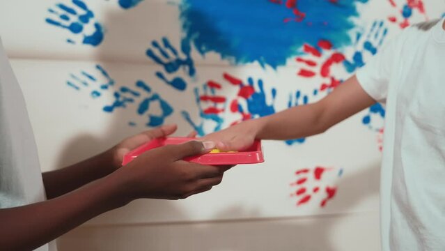 Closeup of happy smart children put hand in color tray and stamp at stained wall. Diverse high school student painting partition with hand print or doing creative activity. Focus on hand. Edification