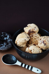 Nut ice cream balls with chocolate in a black bowl and a black and white spoon next to a chocolate donut on a brown background