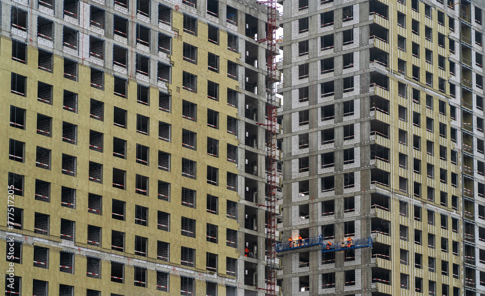 Poster workers on a scaffold install windows on a multistory building clad in yellow insulation