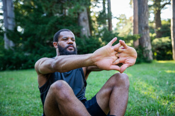 A sporty African man is sitting on the grass and stretching his arms