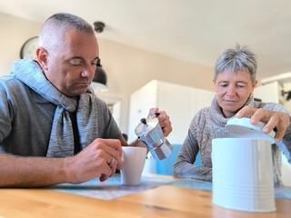 Adult couple, sitting at the table, he seriously pouring the coffee, she with a serene look opening...