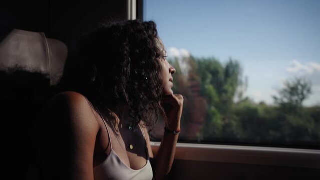 Young African American woman sitting inside train looking pensive and relaxed out the window during the sunset, background field landscape. Traveler millennial people moving in transports in vacation
