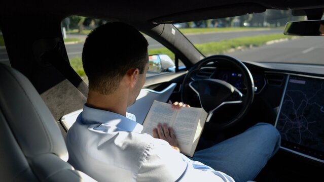 Male businessperson reading book during riding on electrical vehicle with autopilot at urban road. Successful businessman improving his knowledge while riding an autonomous self driving electric car