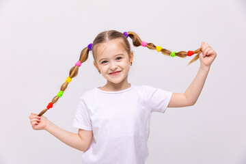 The little girl lifts her braids of hair up, with multi-colored elastic bands, and smiles broadly. Hairstyles for little girls. White isolated background.