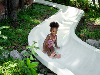 Happy Little African child girl sliding and playing at the swimming pool in the water park on summer vacation.