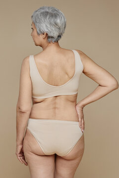 Rear view of senior fit woman in beige lingerie keeping right hand on waist while posing over pastel color background in front of camera