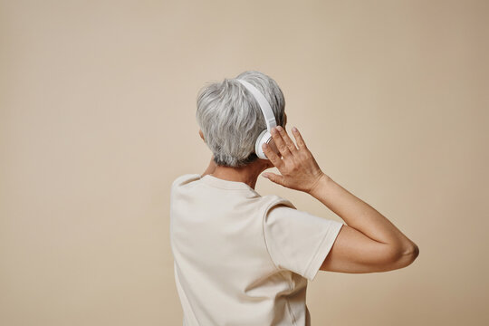 Rear view of senior woman with grey short hair listening to music in headphones while standing on beige background in isolation