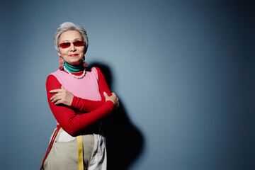 Stylish senior woman in trendy attire crossing arms by chest during photo session while standing by blue wall with copyspace on her left