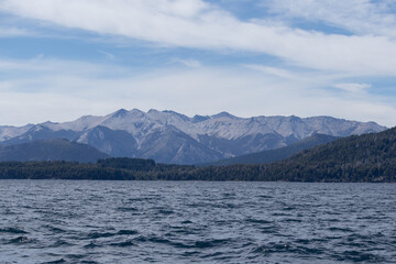 Horizontal photo of mountains full of vegetation and rocks, surrounded by tranquil waves of a lake's water, with a clear blue sky during a sunny day, in Lago Nahuel Huapi, Argentina - Powered by Adobe
