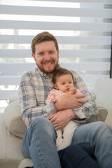 Obraz premium Portrait of dad holding his baby daughter in his arms, It's daytime he looks at the camera sitting on the sofa.