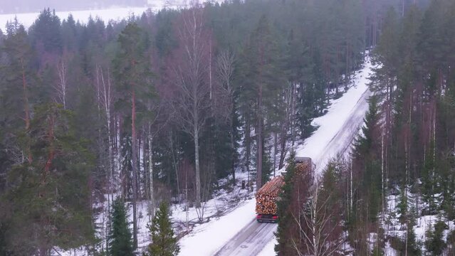 Truck transports harvested timber logs on snowy forest road, telephoto aerial