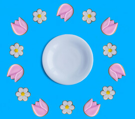Empty white plate for text and gingerbread cookies in the shape of chamomile and tulip on the blue background. Copy space.