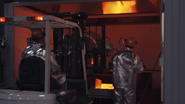 Factory workers in protective heat resistant aluminum foil suits, costumes and helmets at metal cast in the foundry. Man driving forklift truck, loader. Steel making plant, metallurgists. Slow motion