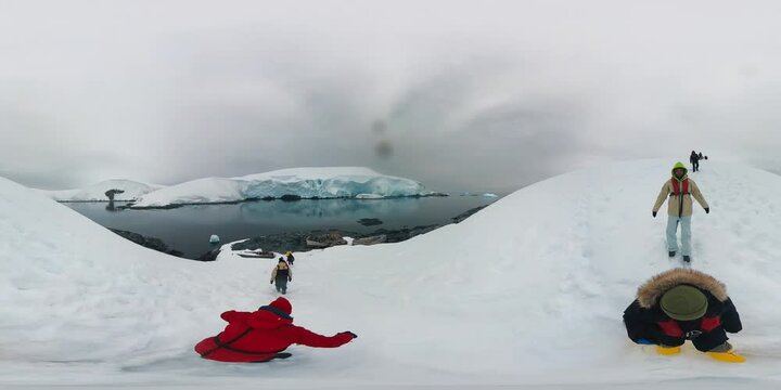 360 VR footage of the tourists walking on the snow in the Antarctica. People in warm jackets walk down the slope in the deserted landscape of the Antarctica and move towards the coast