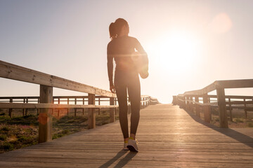 Rear view of a sporty woman carrying an exercise mat, walking along a boardwalk in the sun