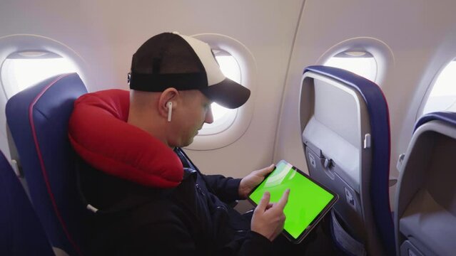 Man with white headphones in ears sitting in airplane holding digital tablet computer with green screen scrolling and sliding with finger on display.